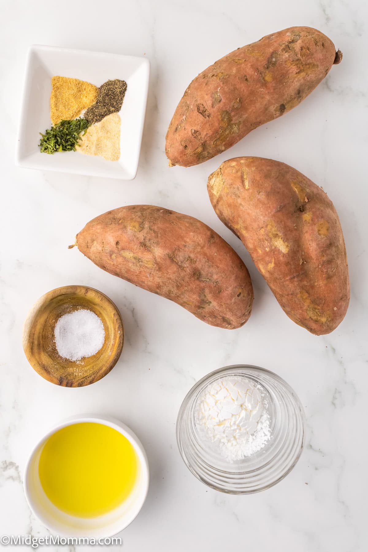 Three raw sweet potatoes on a white surface, accompanied by small bowls of olive oil, salt, cornstarch, and a dish of garlic powder, black pepper, and dried herbs.