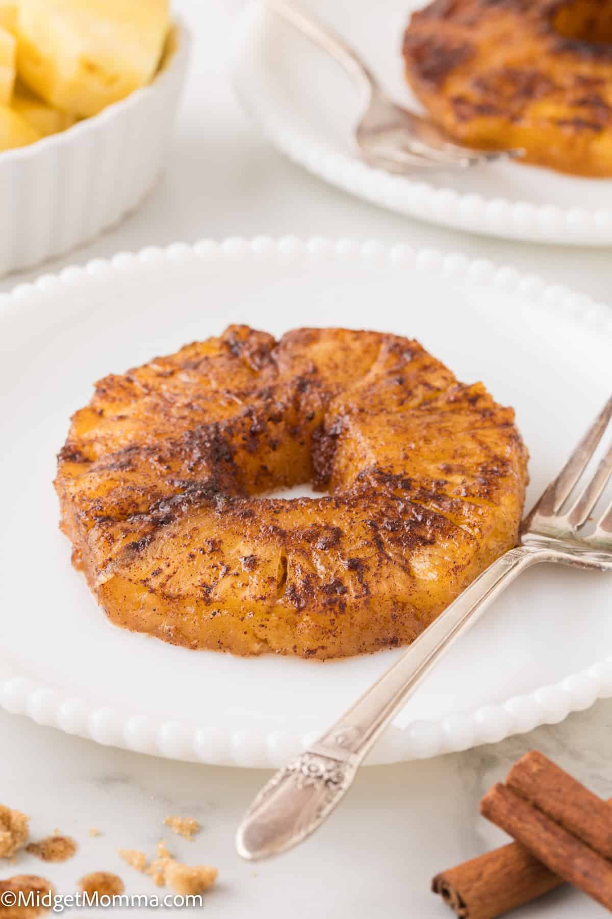 A single baked pineapple ring sprinkled with cinnamon on a white plate, accompanied by a fork and cinnamon sticks.