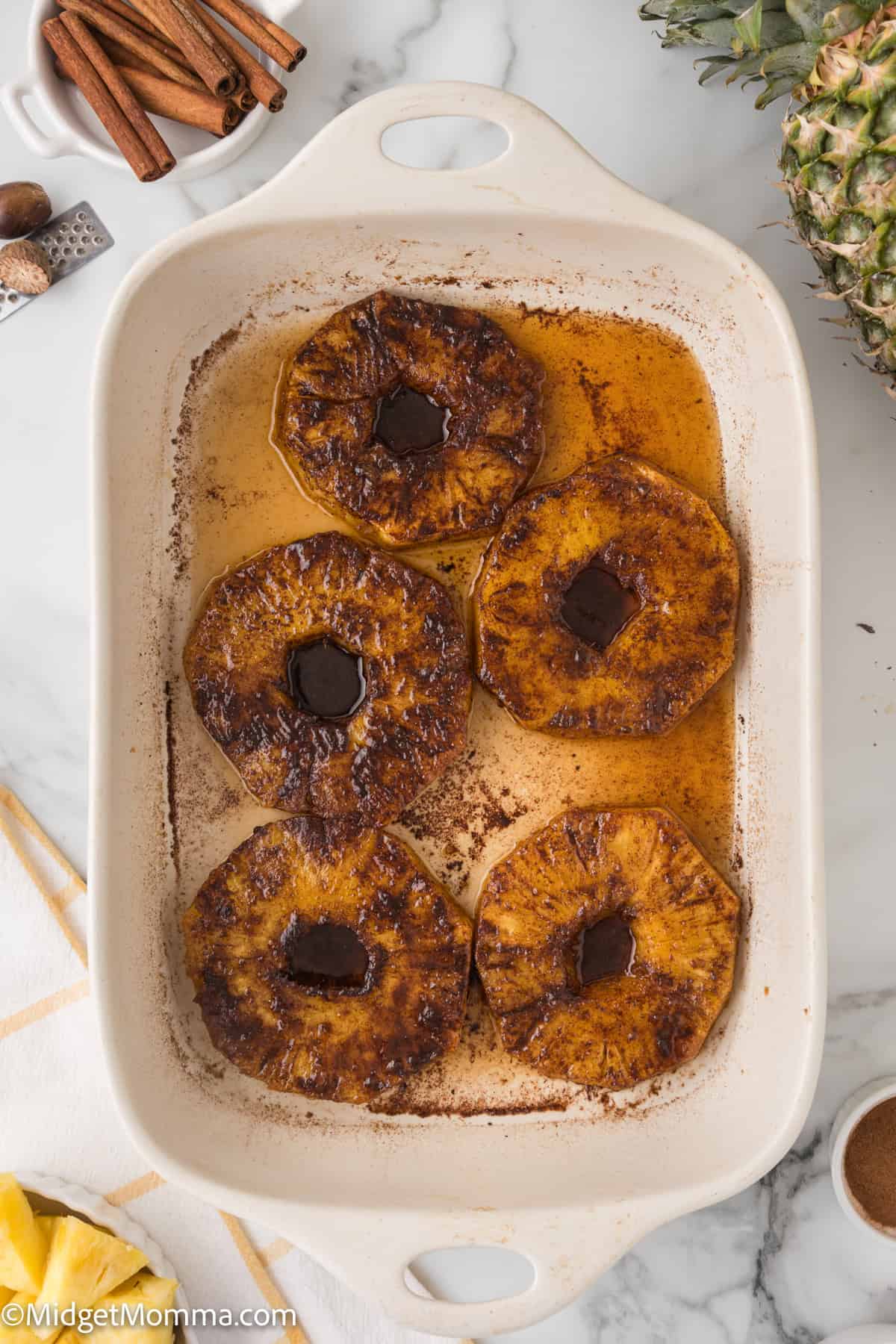Baked pineapple slices with cinnamon in a white baking dish, surrounded by pineapple and cinnamon sticks on a marble countertop.