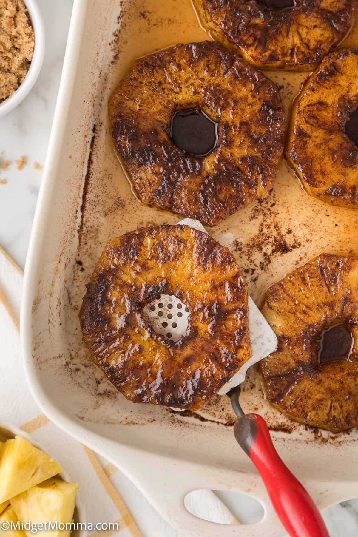 baked pineapple slices in a baking dish, caramelized with a golden-brown char, sprinkled with cinnamon, and a red spatula nearby.