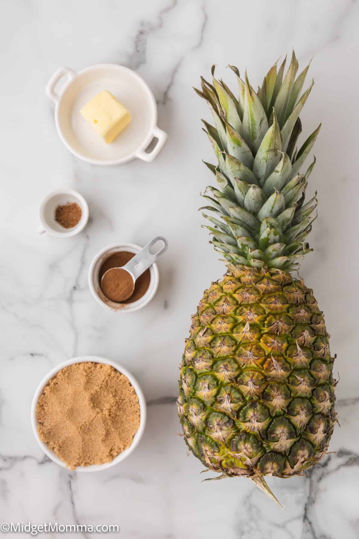 Easy Baked Pineapple Dessert Ingredients. A whole pineapple with ingredients like butter, cinnamon, and brown sugar arranged around it on a marble surface.