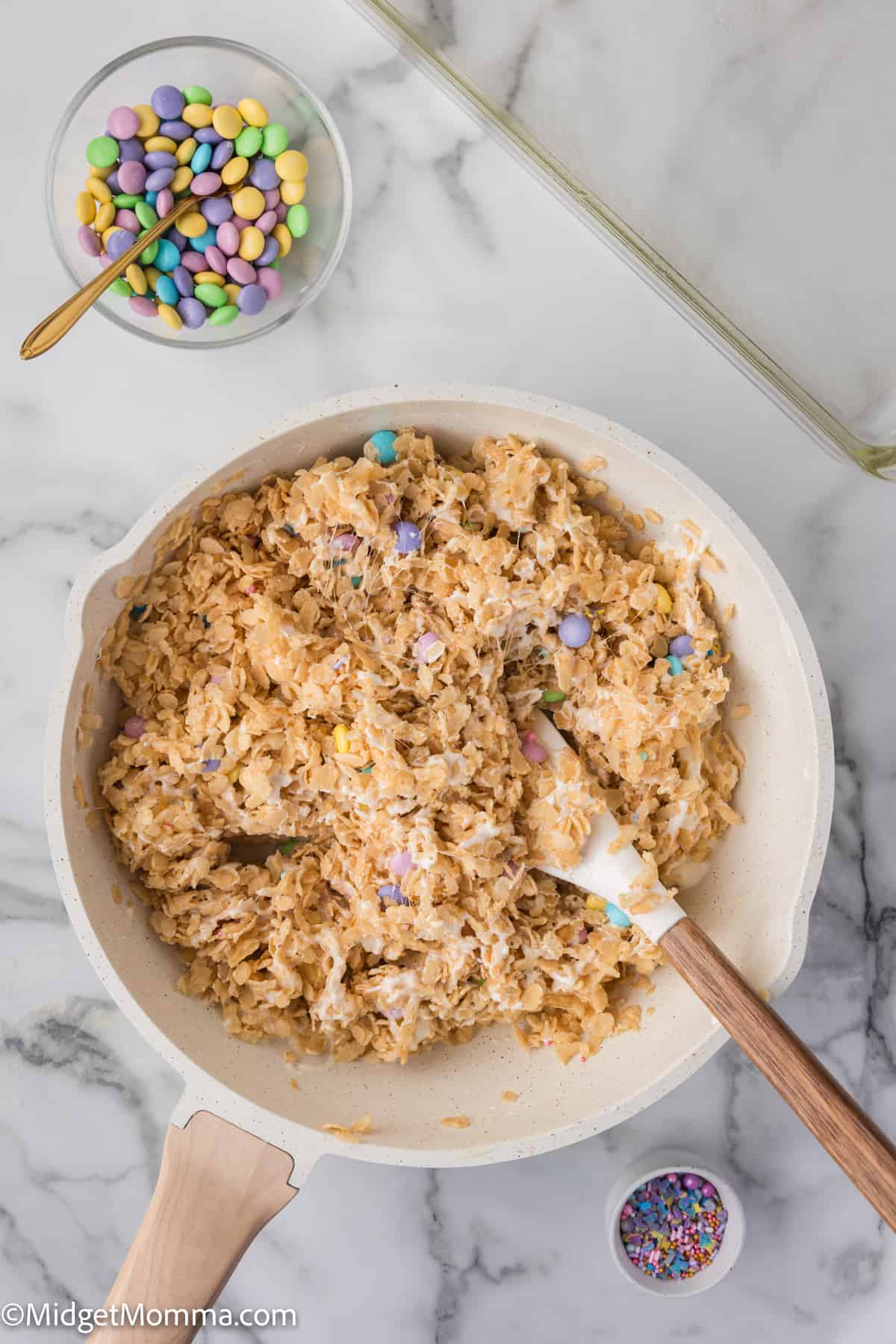 A skillet of rice cereal treats mixed with pastel-colored candies, with extra candies and sprinkles on the side.