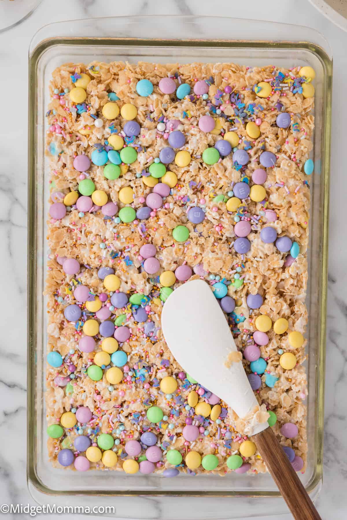 A baking sheet with colorful sprinkles and candy-coated chocolates scattered over marshmallow cereal bars, with a white spatula resting on the surface.