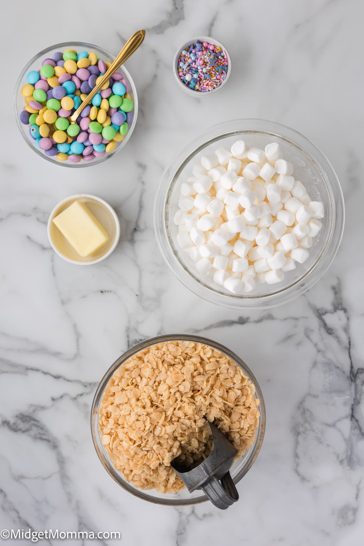 Ingredients for making easter rice krispie treats laid out on a marble countertop, including colorful candies, sprinkles, marshmallows, butter, and crispy rice cereal.