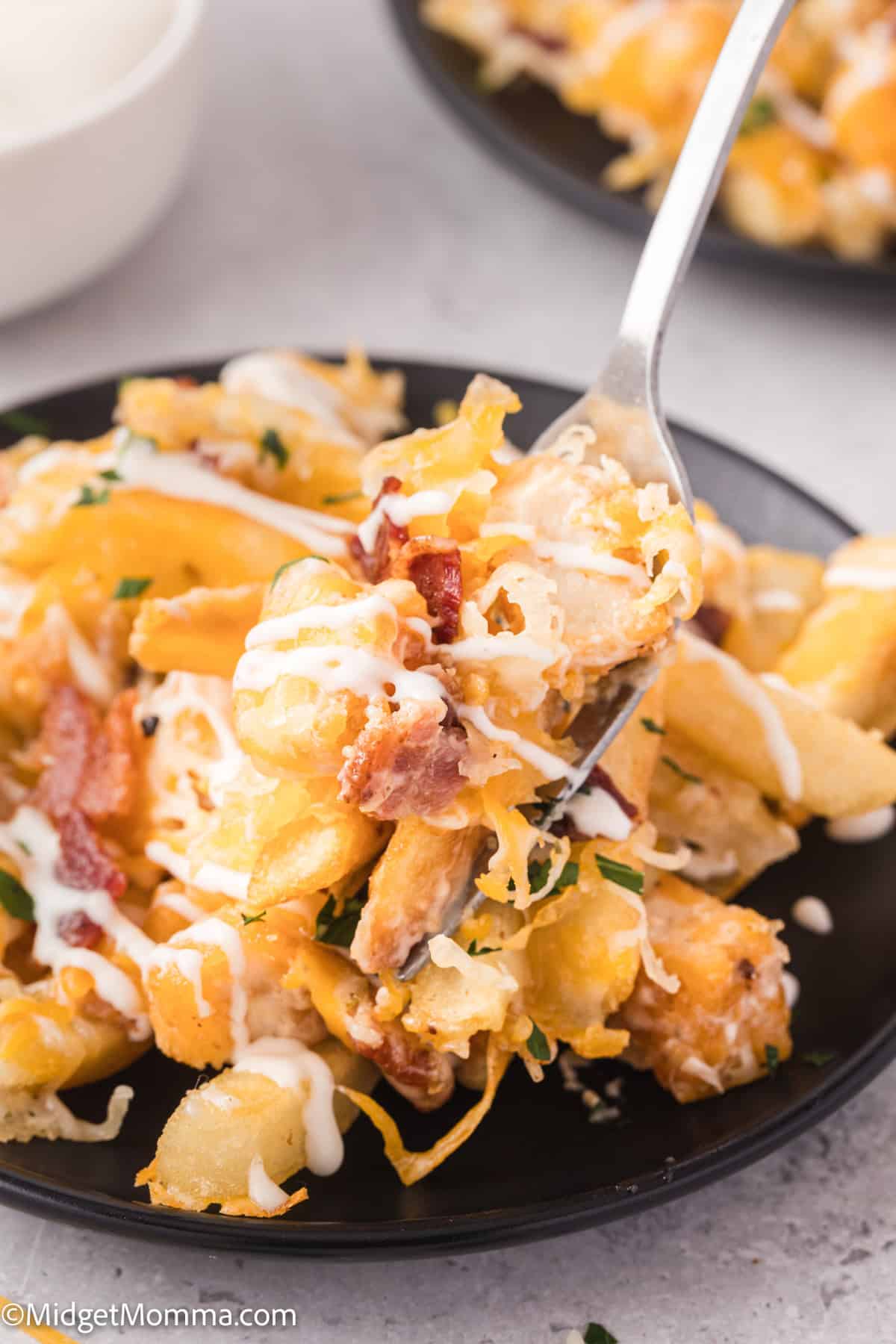 Close-up of a fork holding cheesy loaded fries topped with bacon bits and sauce on a black plate.