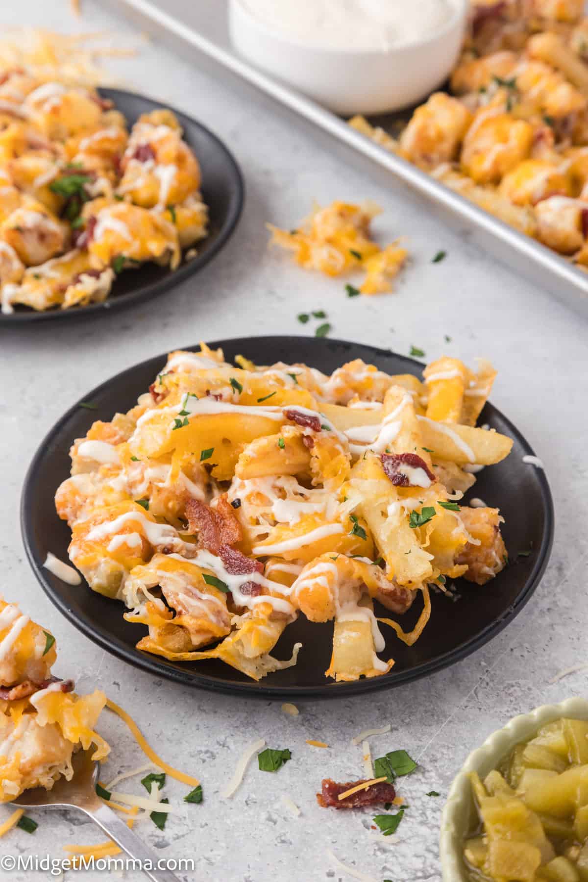 A black plate with cheesy loaded fries topped with bacon and herbs, set on a light surface. Other plates and a bowl of dip are visible in the background.