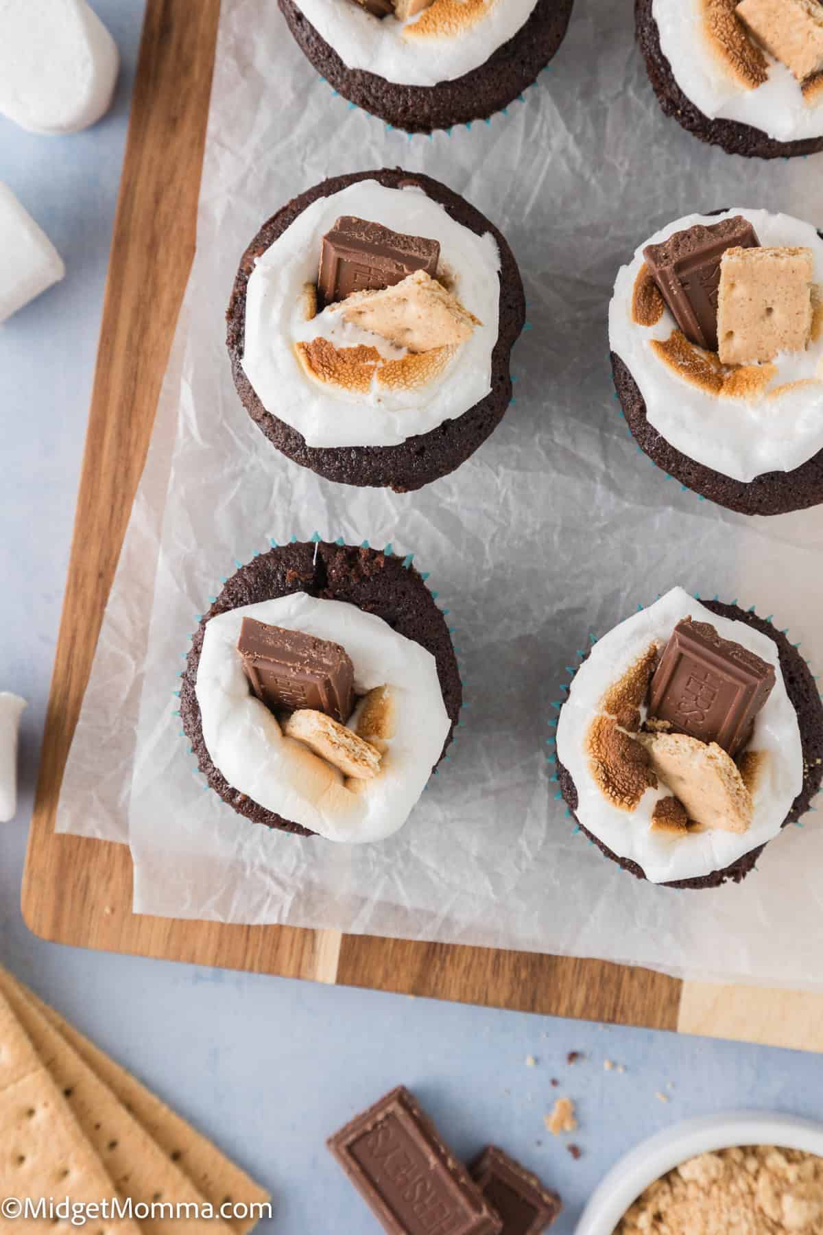 Cupcake s'mores with marshmallow topping and chocolate pieces on a wooden board, surrounded by graham crackers and marshmallows.