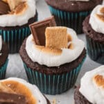 Chocolate cupcakes topped with toasted marshmallows, a piece of chocolate, and a graham cracker in teal cupcake liners.