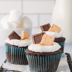 Three s'mores cupcakes with a melted marshmallow, topped with a piece of chocolate and a graham cracker, on a cooling rack.