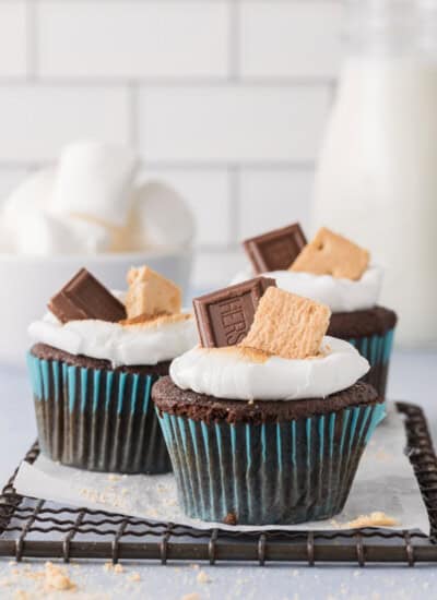 Three s'mores cupcakes with a melted marshmallow, topped with a piece of chocolate and a graham cracker, on a cooling rack.