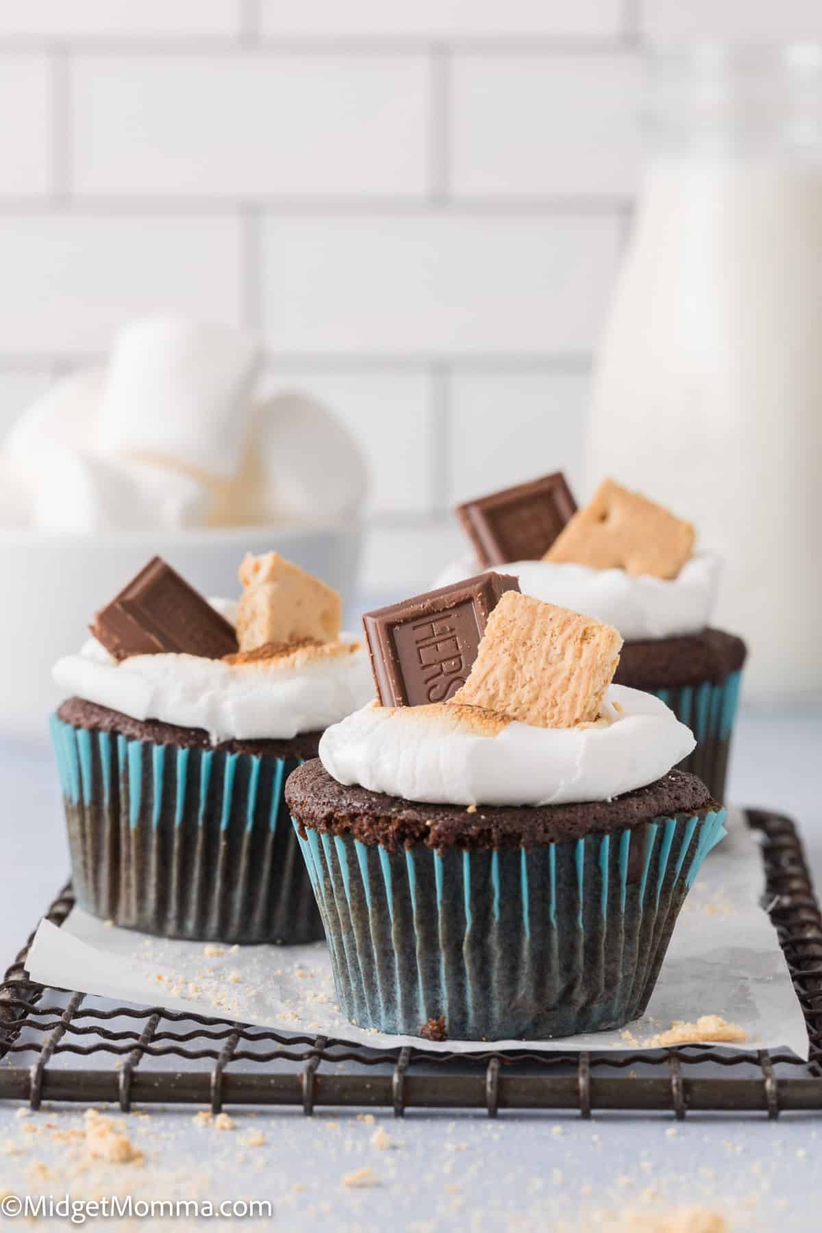 Three s'mores cupcakes with a melted marshmallow, topped with a piece of chocolate and a graham cracker, on a cooling rack.