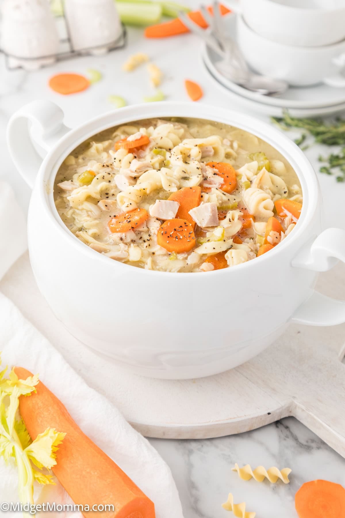 A white soup tureen filled with chicken noodle soup containing carrots, celery, chicken pieces, and pasta sits on a marble surface with vegetables and herbs nearby.