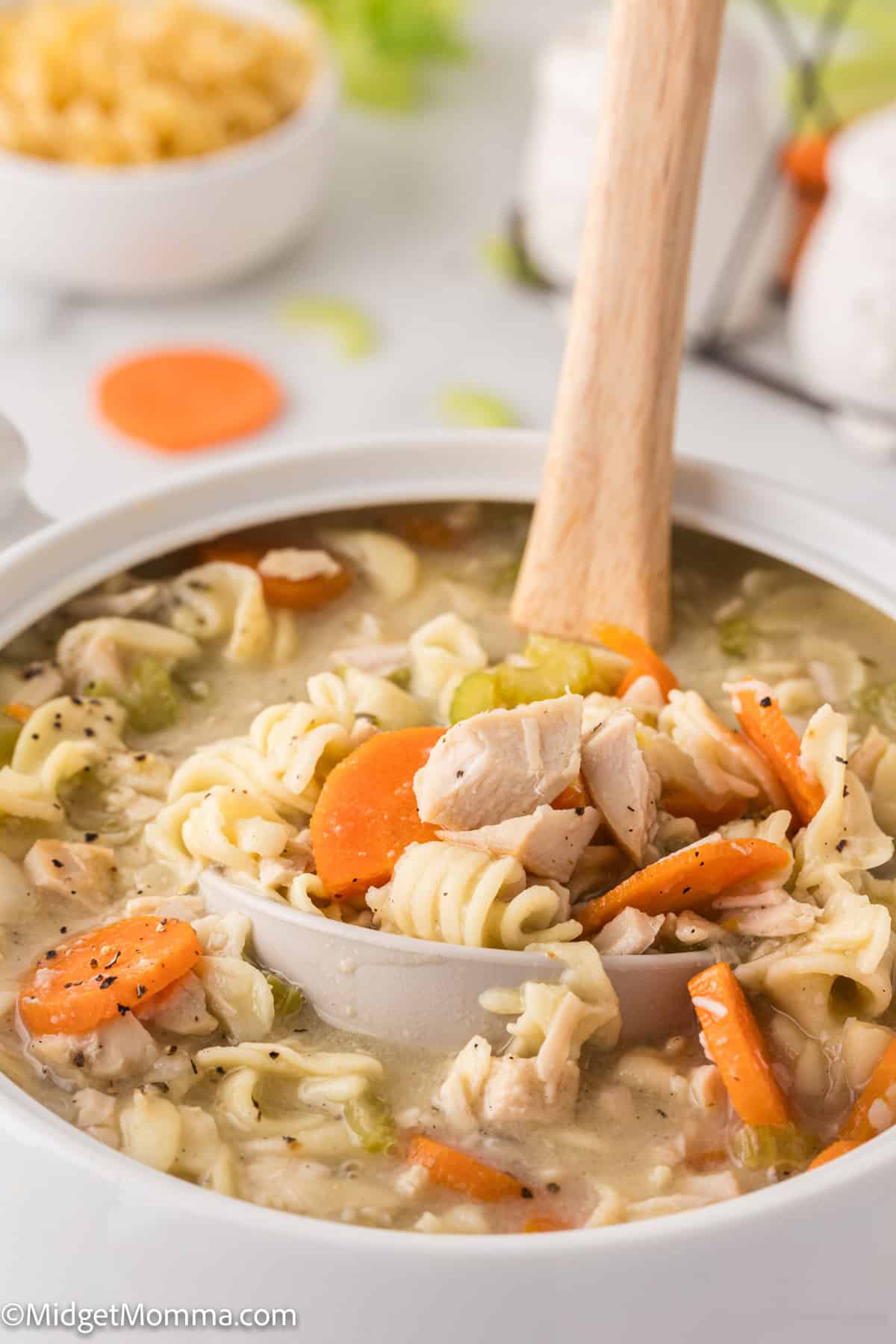 A ladle filled with chicken noodle soup containing rotini pasta, carrots, celery, and chicken pieces over a white pot.