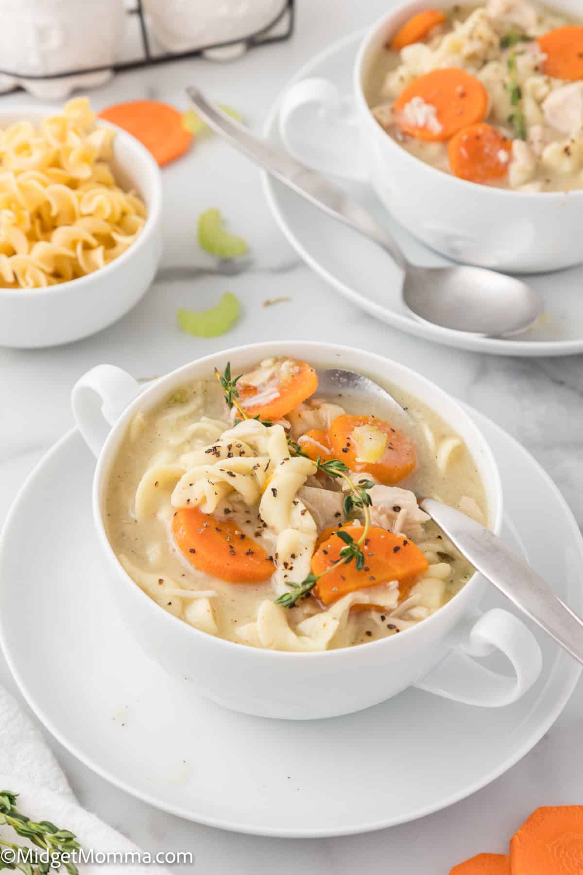 A bowl of chicken noodle soup with carrots, noodles, and herbs, served on a white plate with a spoon. Another bowl and a small dish of noodles are in the background.