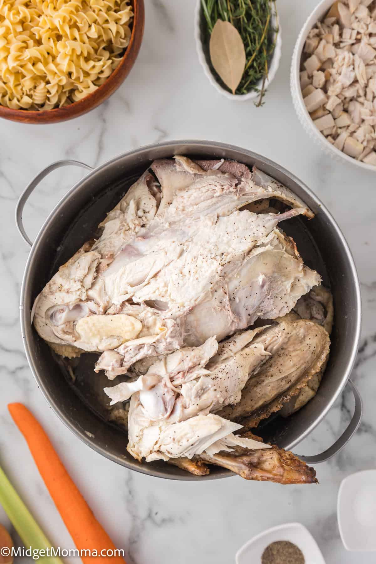 A large pot contains a cooked turkey carcass, surrounded by chopped vegetables, a bowl of diced turkey, herbs, and uncooked noodles on a marble countertop.