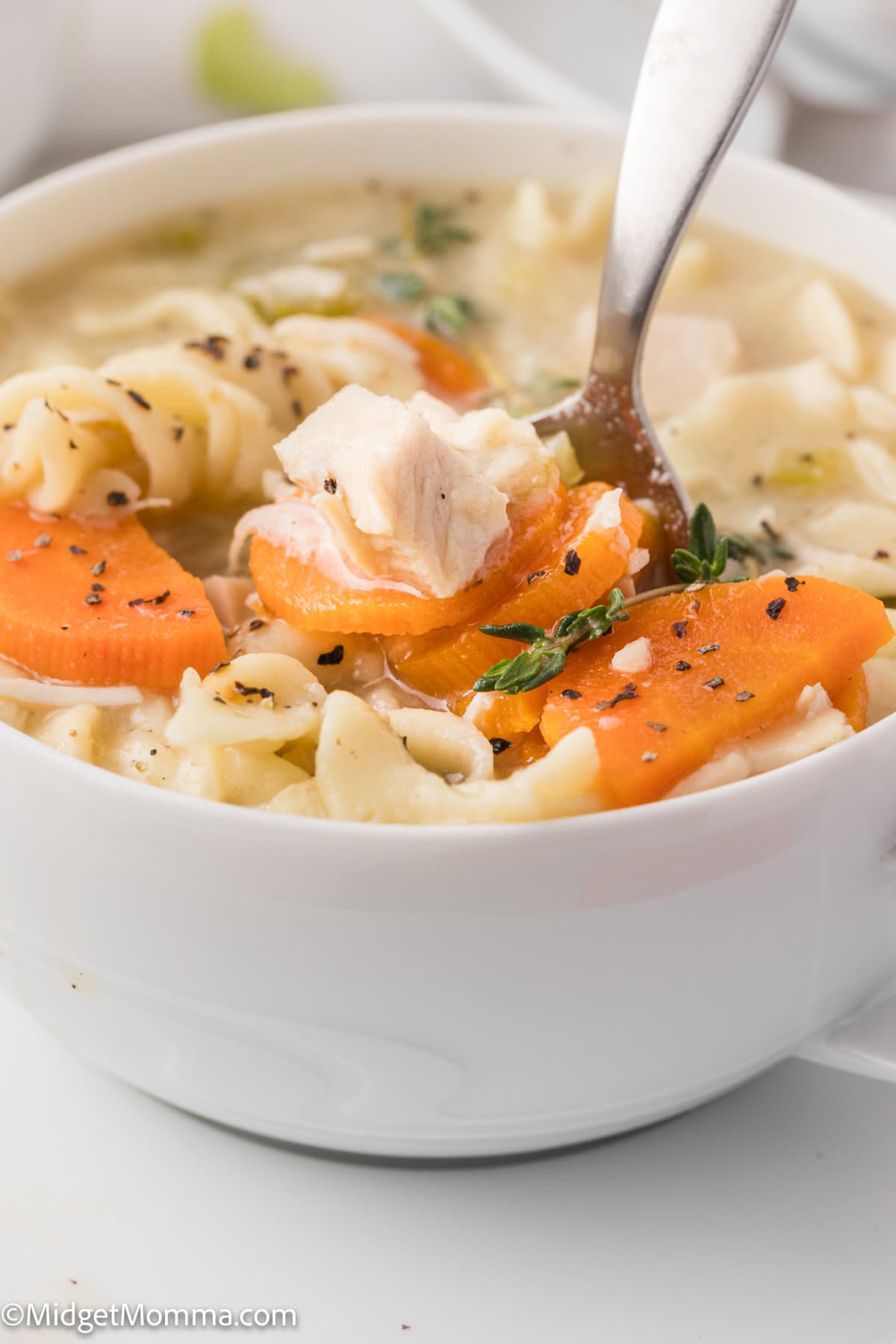 A bowl of chicken noodle soup with carrots, pasta, and herbs, with a spoon lifting a bite.