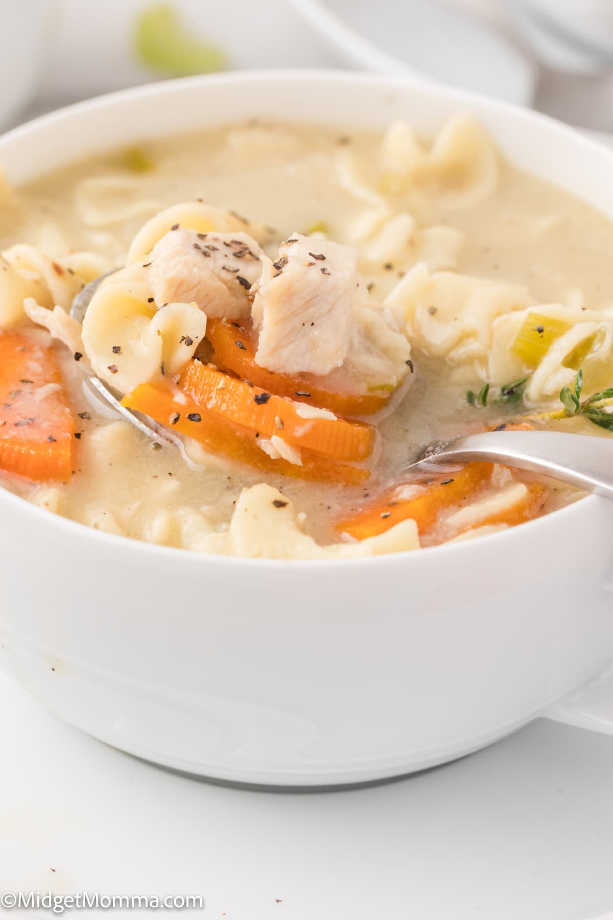 A close-up of a bowl of chicken noodle soup with carrots, noodles, and herbs, with a spoon inside the bowl.