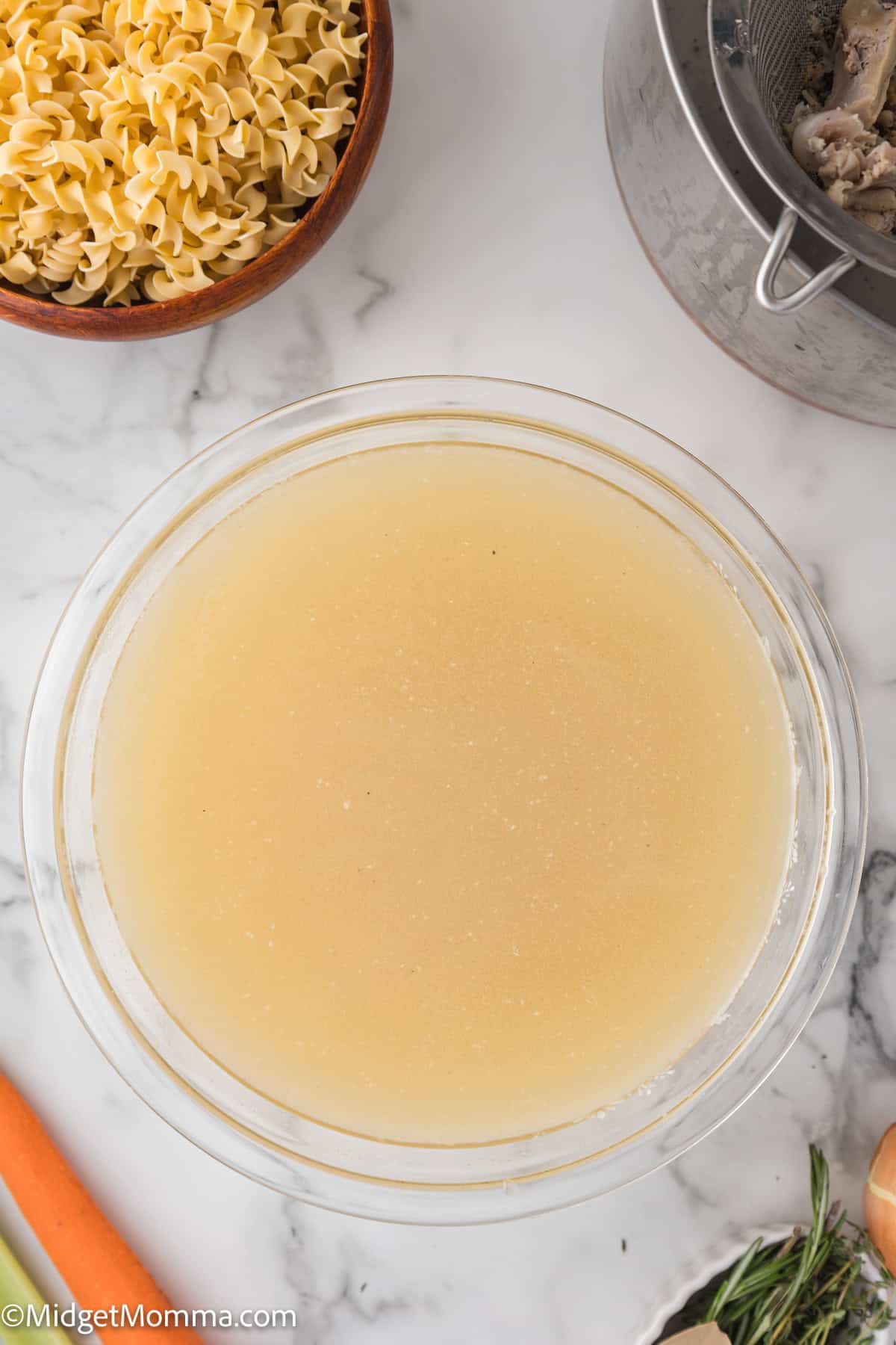 A glass bowl filled with light-colored broth sits on a marble countertop near a bowl of egg noodles, a metal pot, a carrot, and celery.