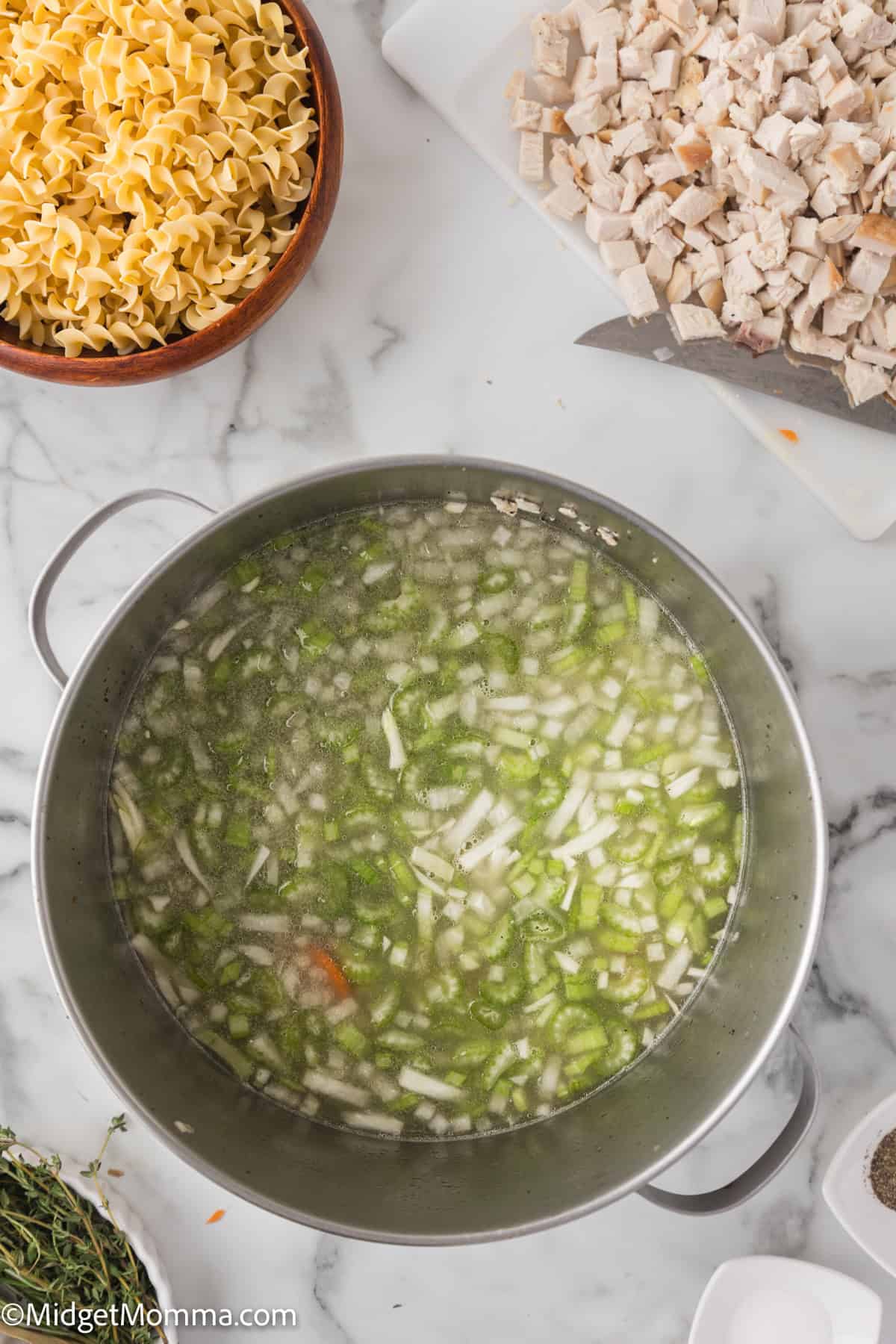 A pot of soup with diced chicken, celery, and onions sits on a marble countertop next to a bowl of uncooked egg noodles and a cutting board with a knife.