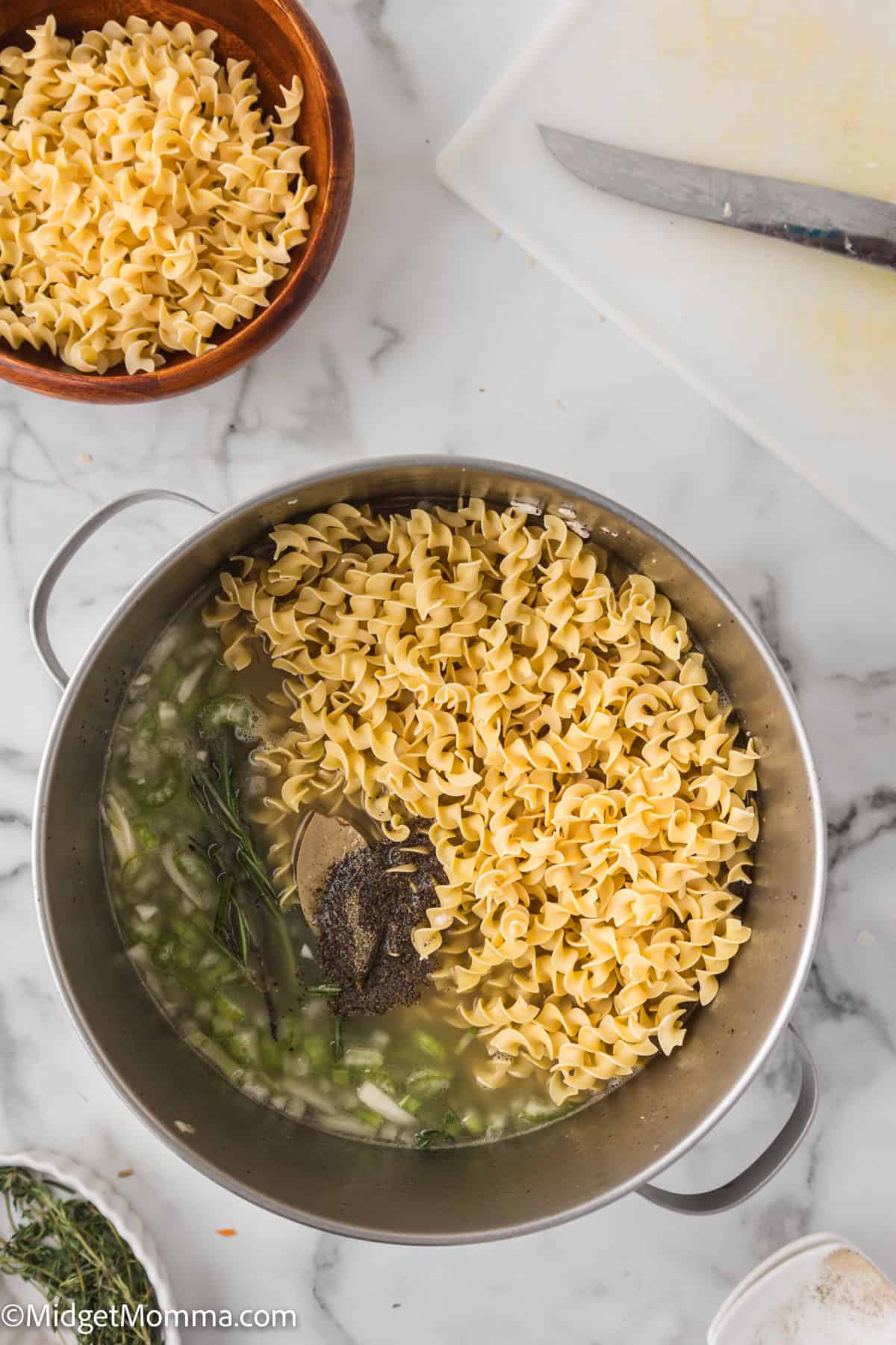 A pot with uncooked egg noodles, herbs, and chopped vegetables in water sits on a marble countertop near a bowl of noodles, a knife, and a cutting board.