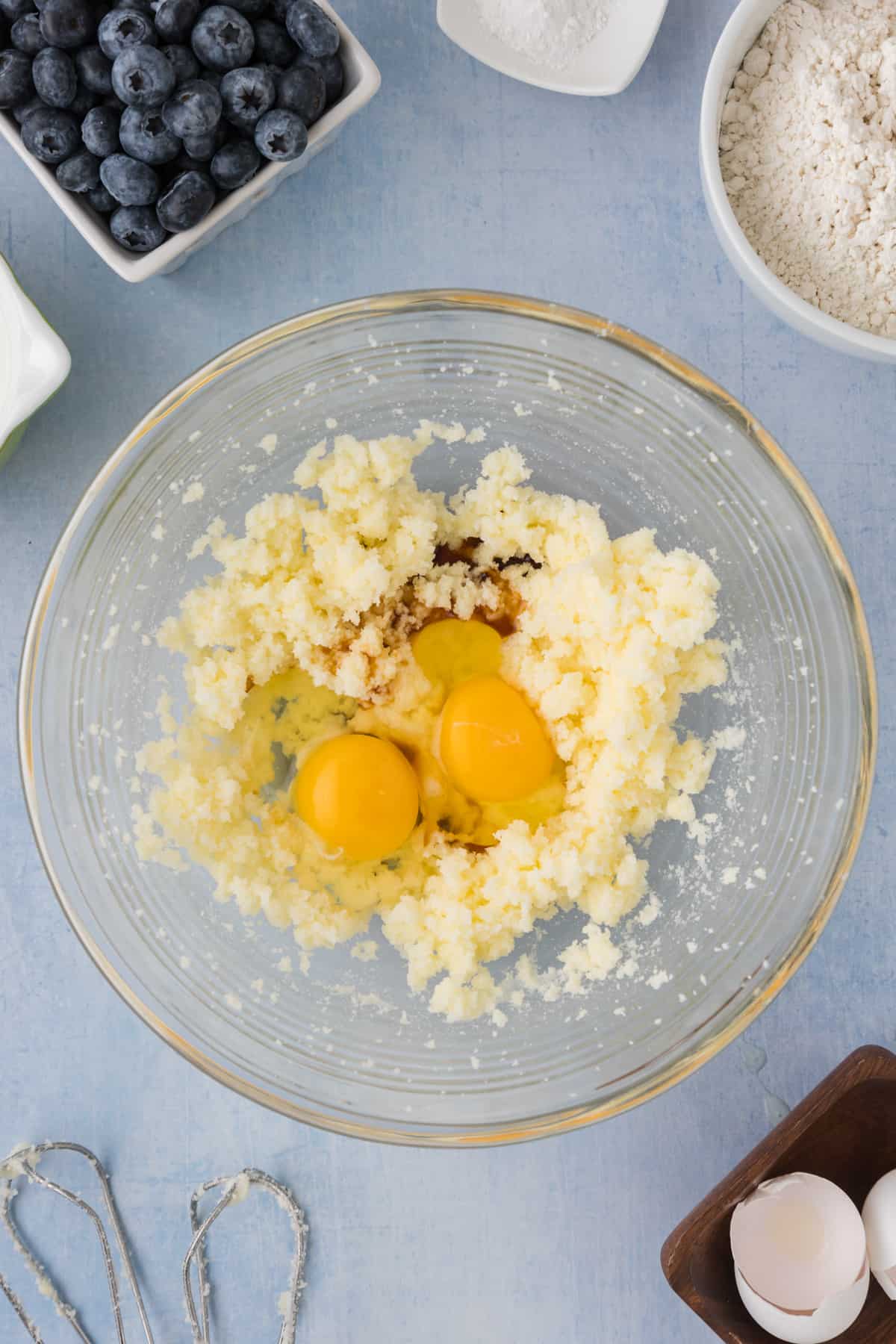 Eggs, flour, sugar and blueberries in a bowl on a blue background.