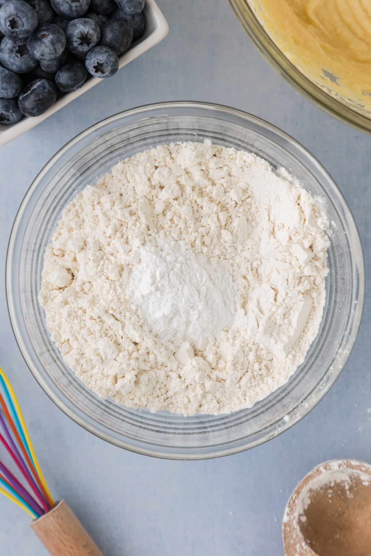 A bowl of flour and blueberries next to a whisk.
