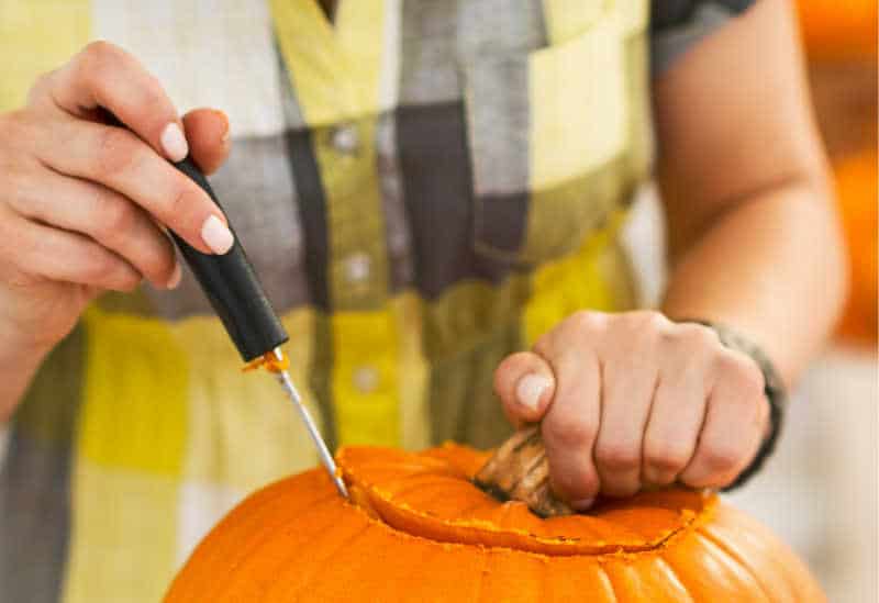 Closeup on person in a Halloween decorated kitchen carving a big orange pumpkin Jack-O-Lantern