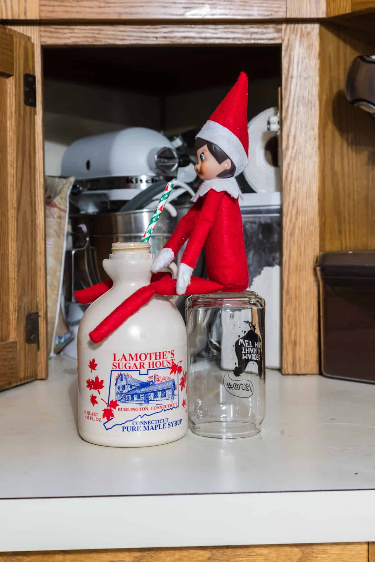 The elf on the shelf is sitting on top of a jar drinking maple syrup with a straw from the jug of maple syrup