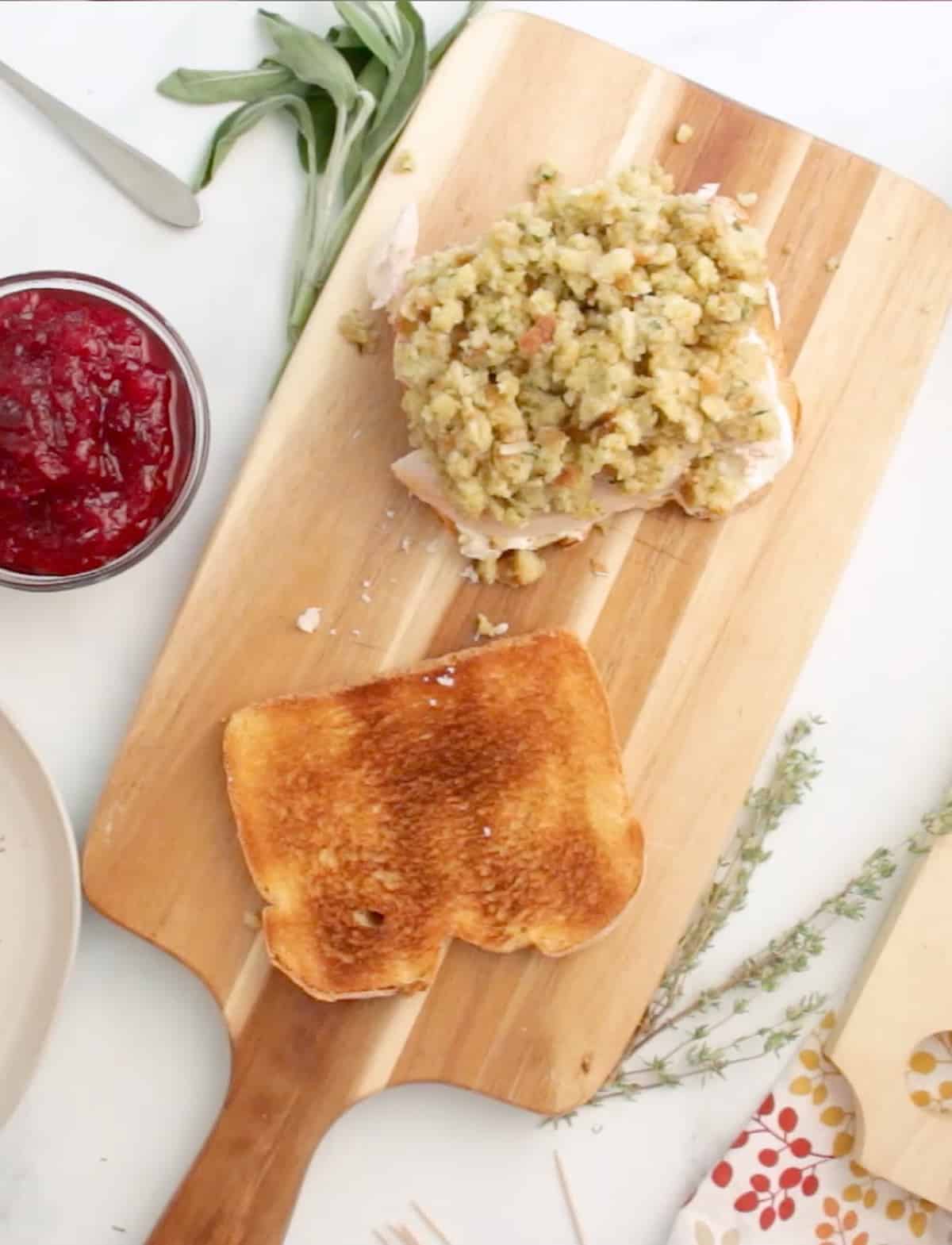 A wooden cutting board with turkey gobbler sandwich being made - stuffing layer being added and cranberry sauce in a bowl.
