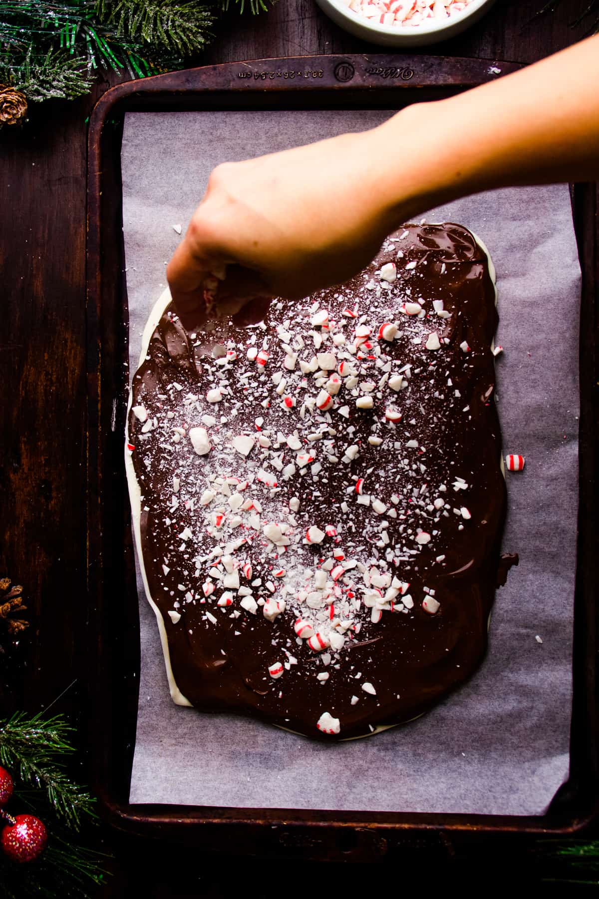 A person is sprinkling crushed peppermint on top of peppermint chocolate on a baking sheet.