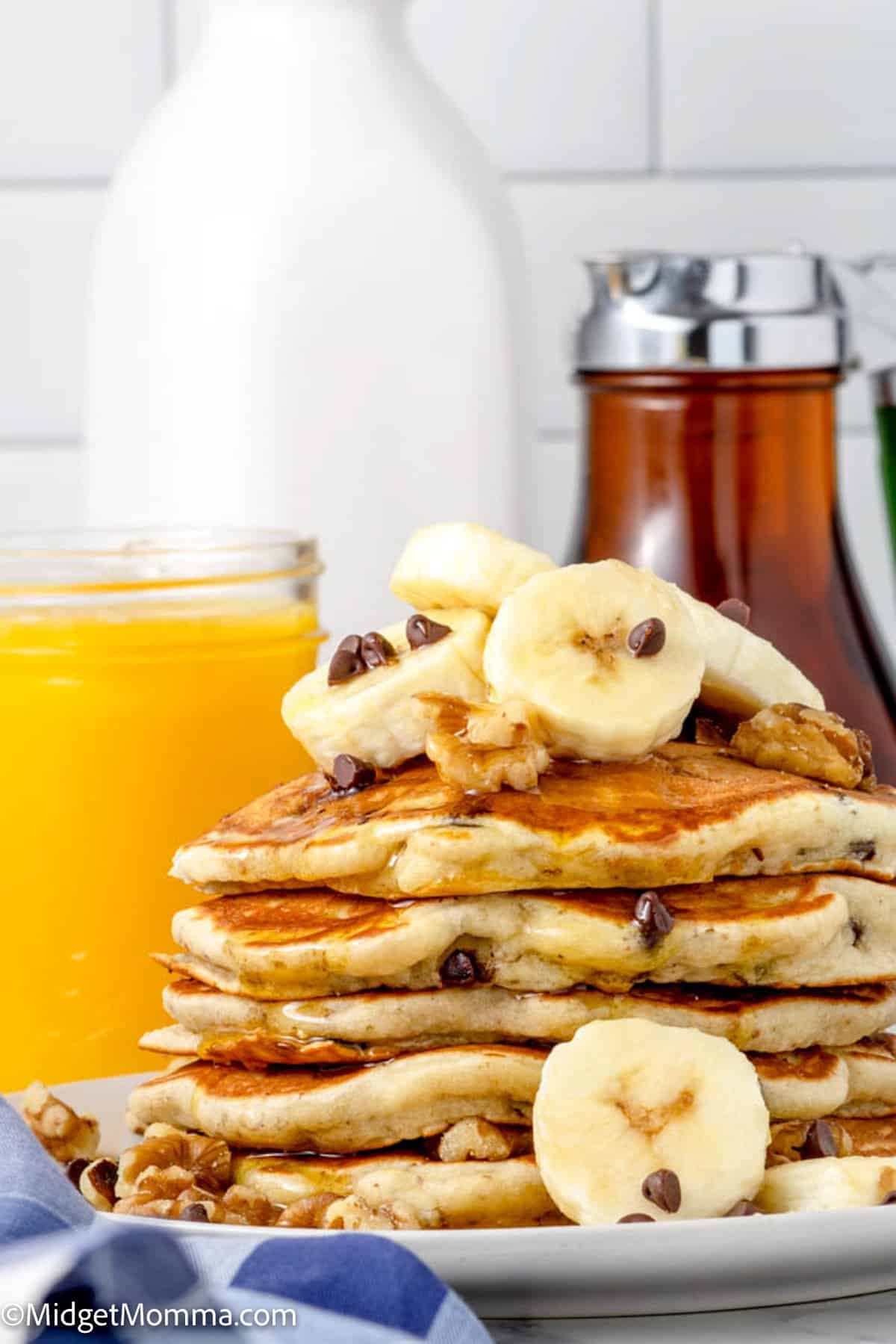 close up photo of a stack Chunky Monkey Pancakes topped with banana slices, chocolate chips and walnuts on a plate with a cup of orange juice in the background and a bottle of real maple syrup 