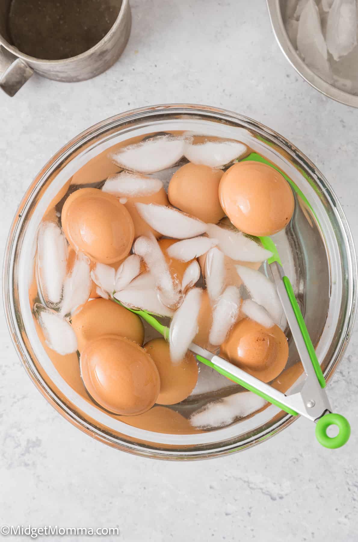 Eggs in a bowl with ice and water and green tongs.