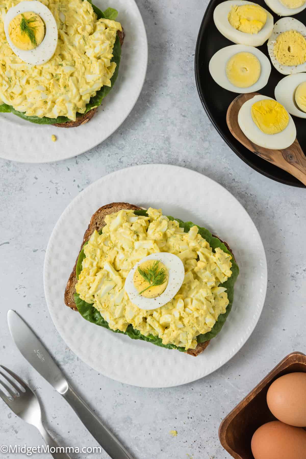 Egg salad served on whole grain bread, topped with egg slices, accompanied by a spoonful of eggs and a carton of brown eggs on a light gray surface.