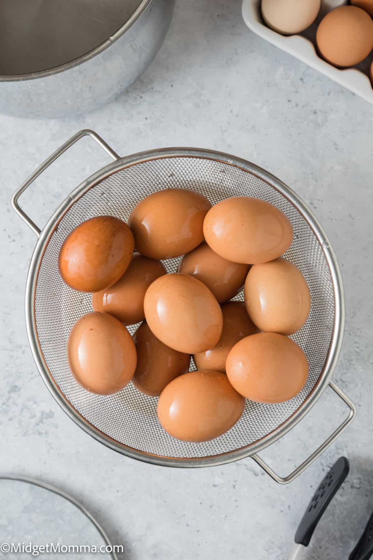 Brown eggs in a metal strainer on a kitchen counter, with a pot and additional eggs in the background.