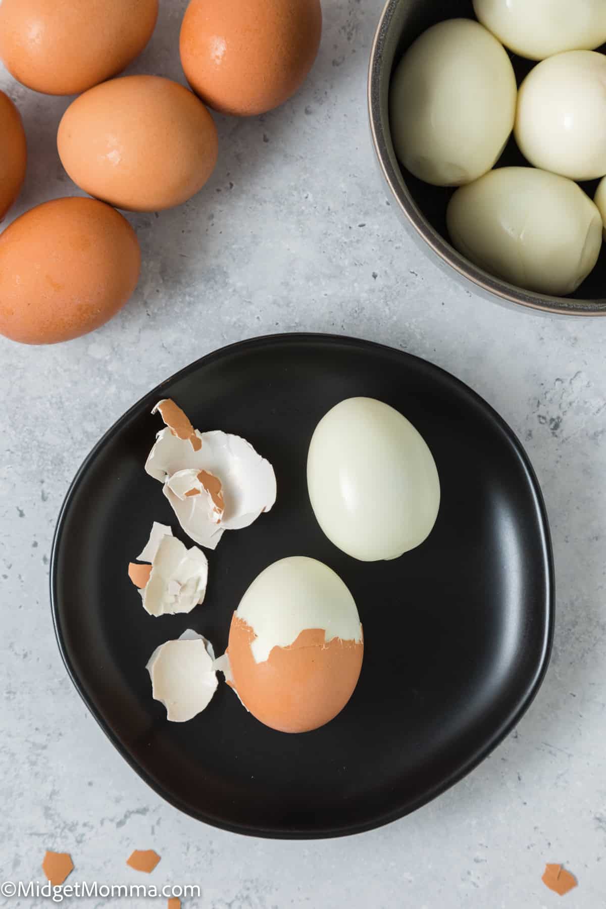 A black plate with one whole peeled hard-boiled egg and eggshell fragments, beside a bowl of unpeeled eggs and a bowl of peeled eggs on a light gray surface.