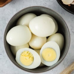A bowl of hard-boiled eggs, some whole and one cut in half, showing the solid yellow yolk, placed on a light surface.