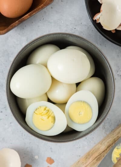 A bowl of hard-boiled eggs, some whole and one cut in half, showing the solid yellow yolk, placed on a light surface.