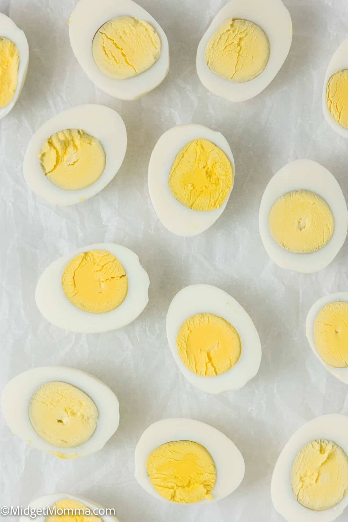 Halved hard-boiled eggs arranged neatly on a crinkled white paper background, showing smooth white exteriors and solid yellow yolks.