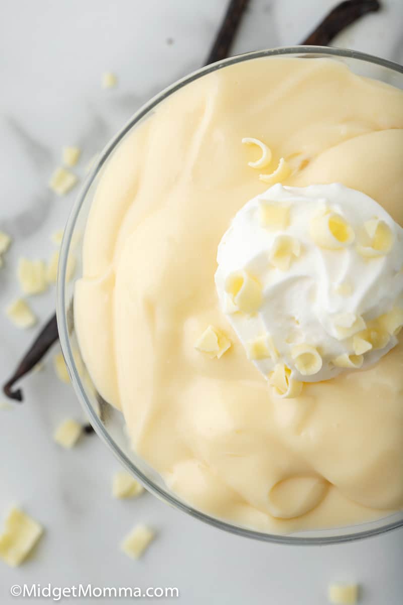 overhead photo of homemade vanilla pudding in a servinig bowl topped with whipped cream and white chocolate shavinigs