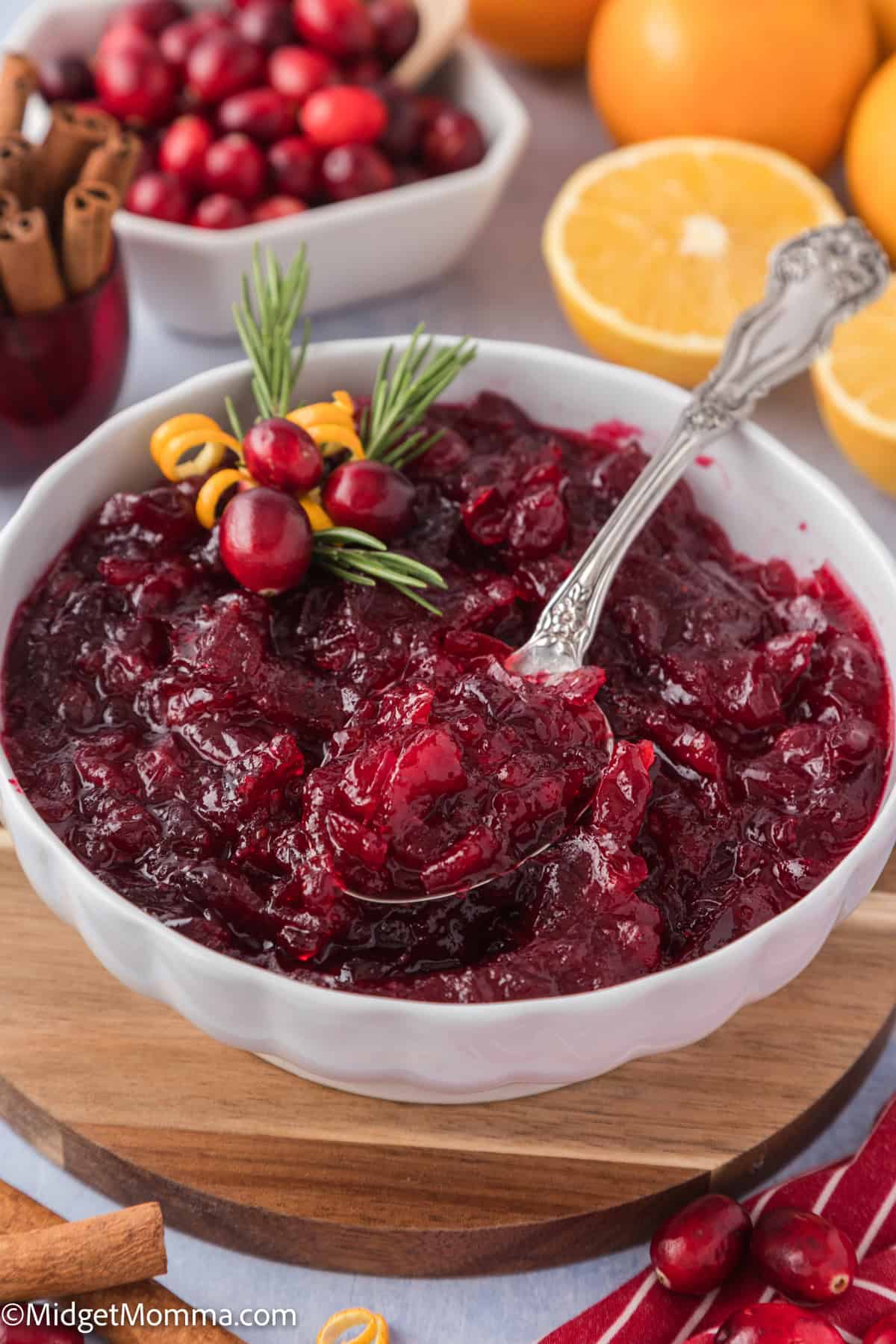Bowl of cranberry sauce garnished with rosemary and cranberries, with a spoon inside. Orange slices and additional cranberries are visible in the background.