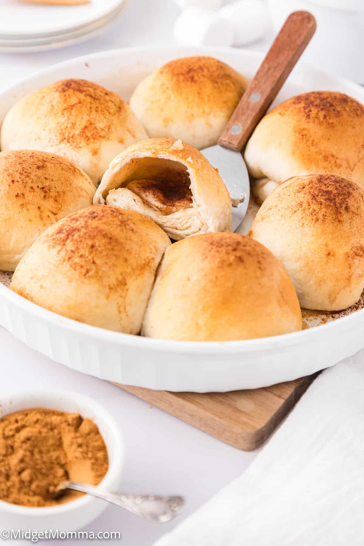 A round white dish with eight baked rolls, one partially opened, revealing filling. A serving spatula is beneath the open roll. A small bowl of spice mix is nearby.