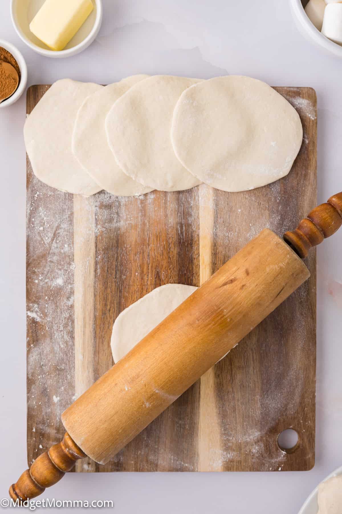 Wooden rolling pin and rolled dough on a cutting board, surrounded by small bowls containing butter and spices.
