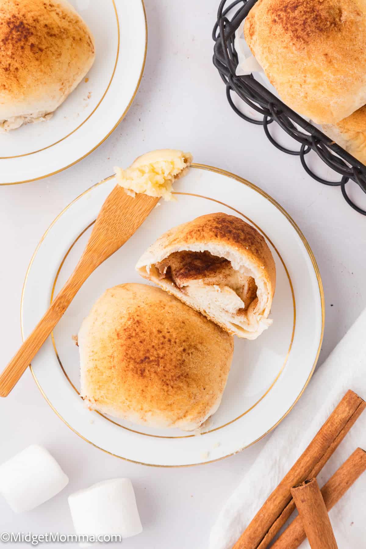 Two cinnamon sugar rolls on a plate, one split open. A wooden spreader with butter rests on the plate. Nearby are more rolls in a basket, cinnamon sticks, and marshmallows.