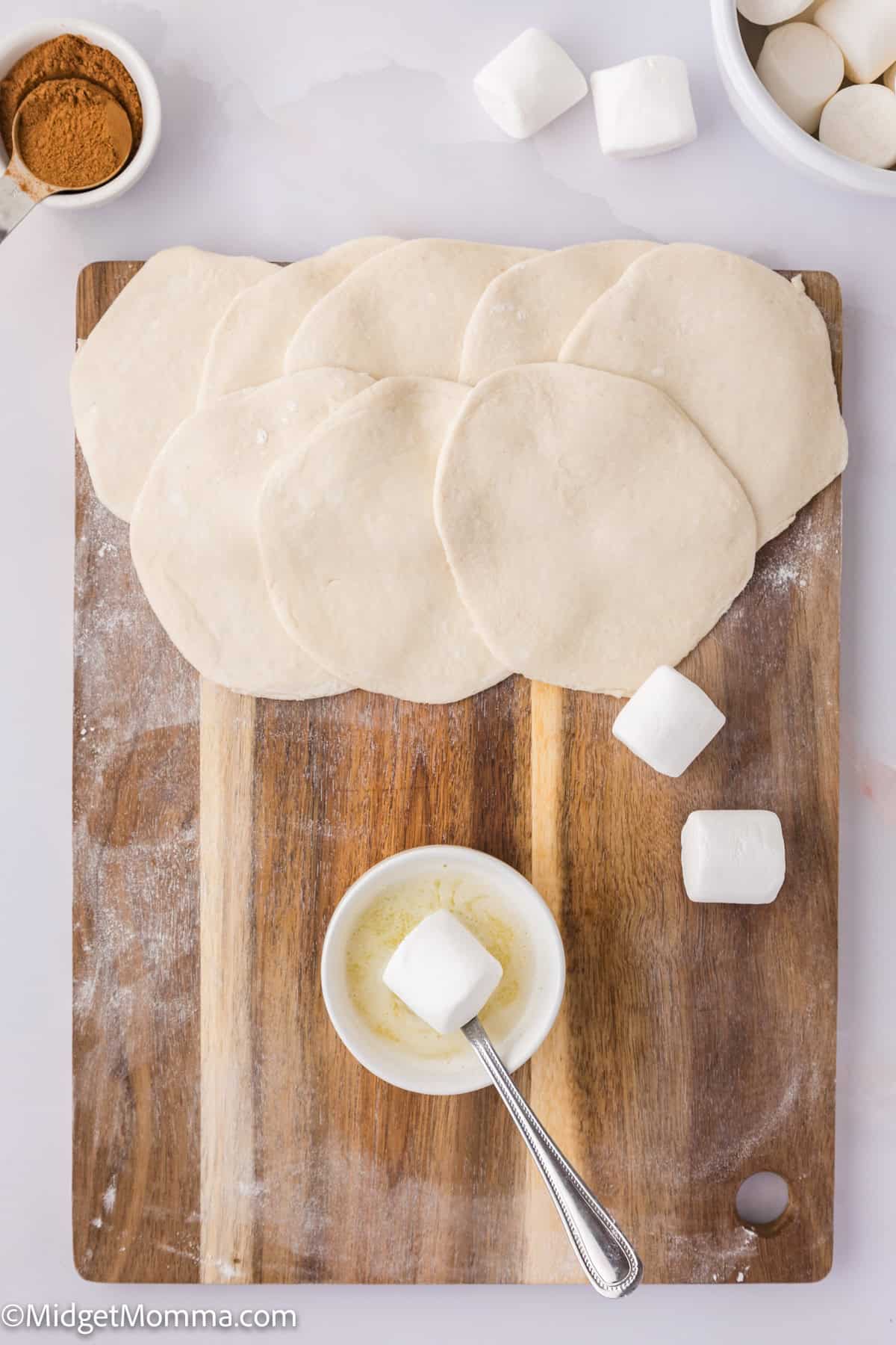 Dough pieces on a wooden board with marshmallows, a bowl of butter, and cinnamon in the background.