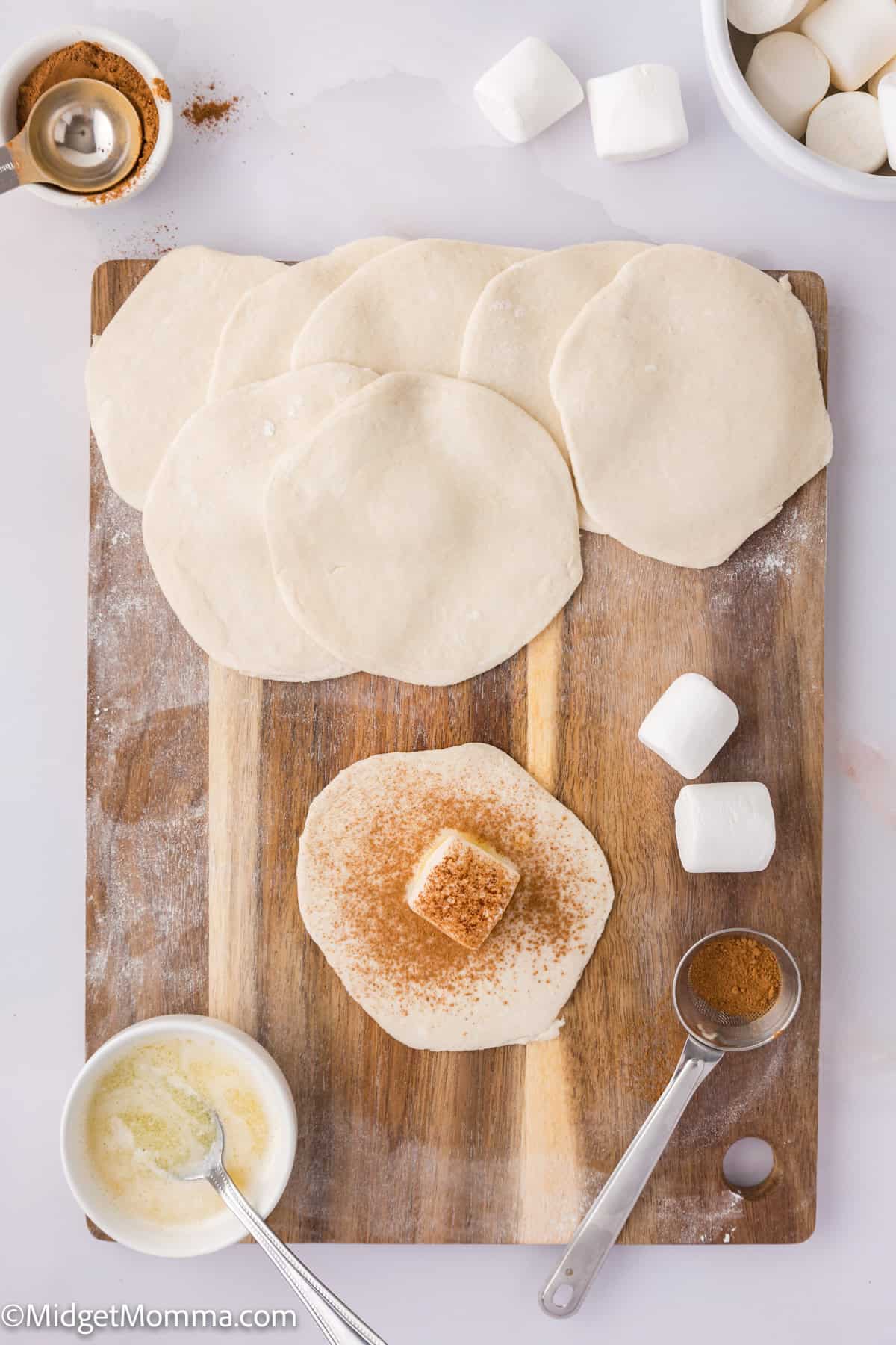 Wooden board with raw dough circles, one topped with a marshmallow and cinnamon. Nearby are more marshmallows, a bowl of flour, and a spoon with cinnamon.