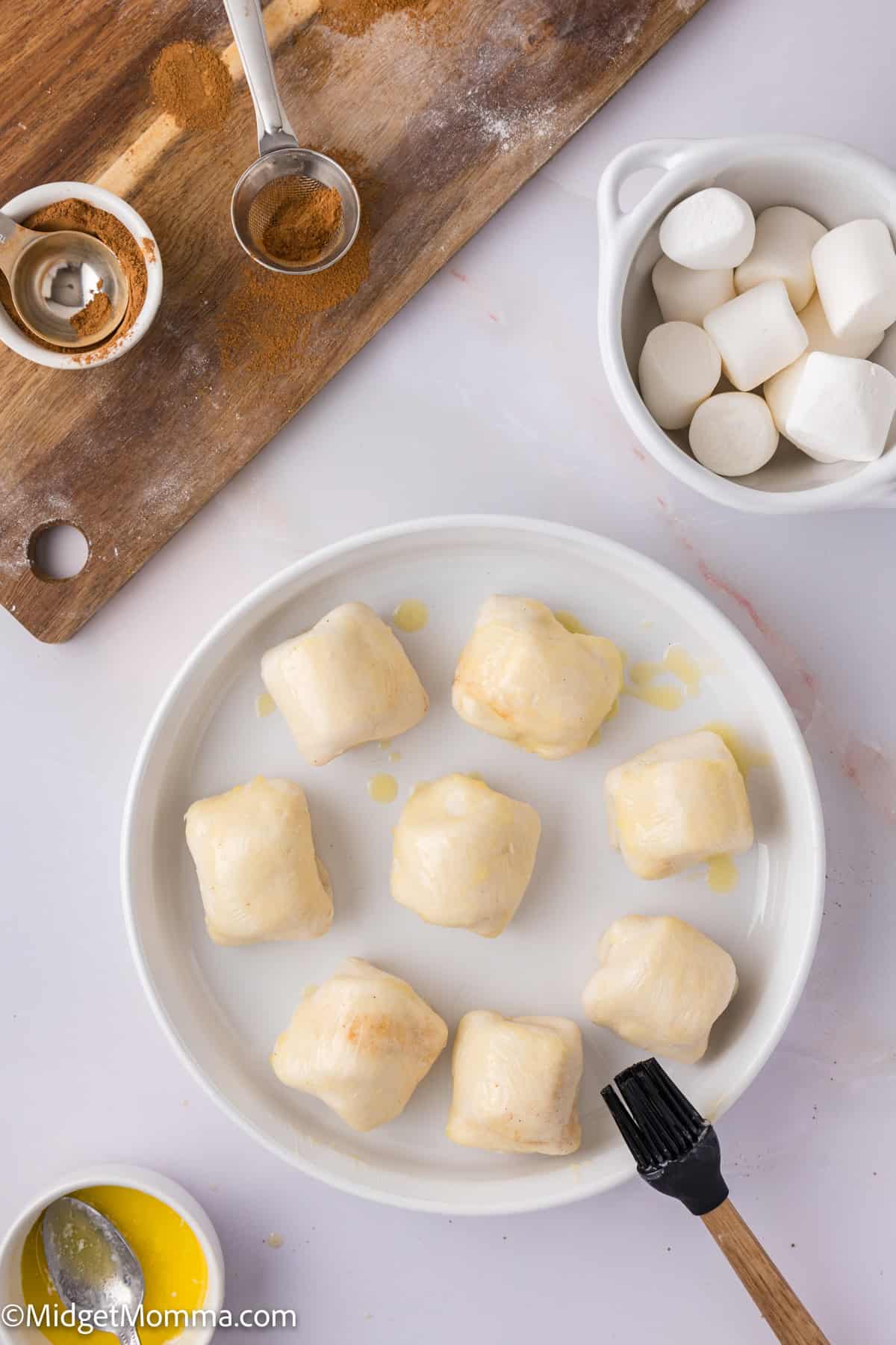 Unbaked pastry dough balls on a white plate with a brush, near a bowl of marshmallows and a wooden board with cinnamon and a spoon.