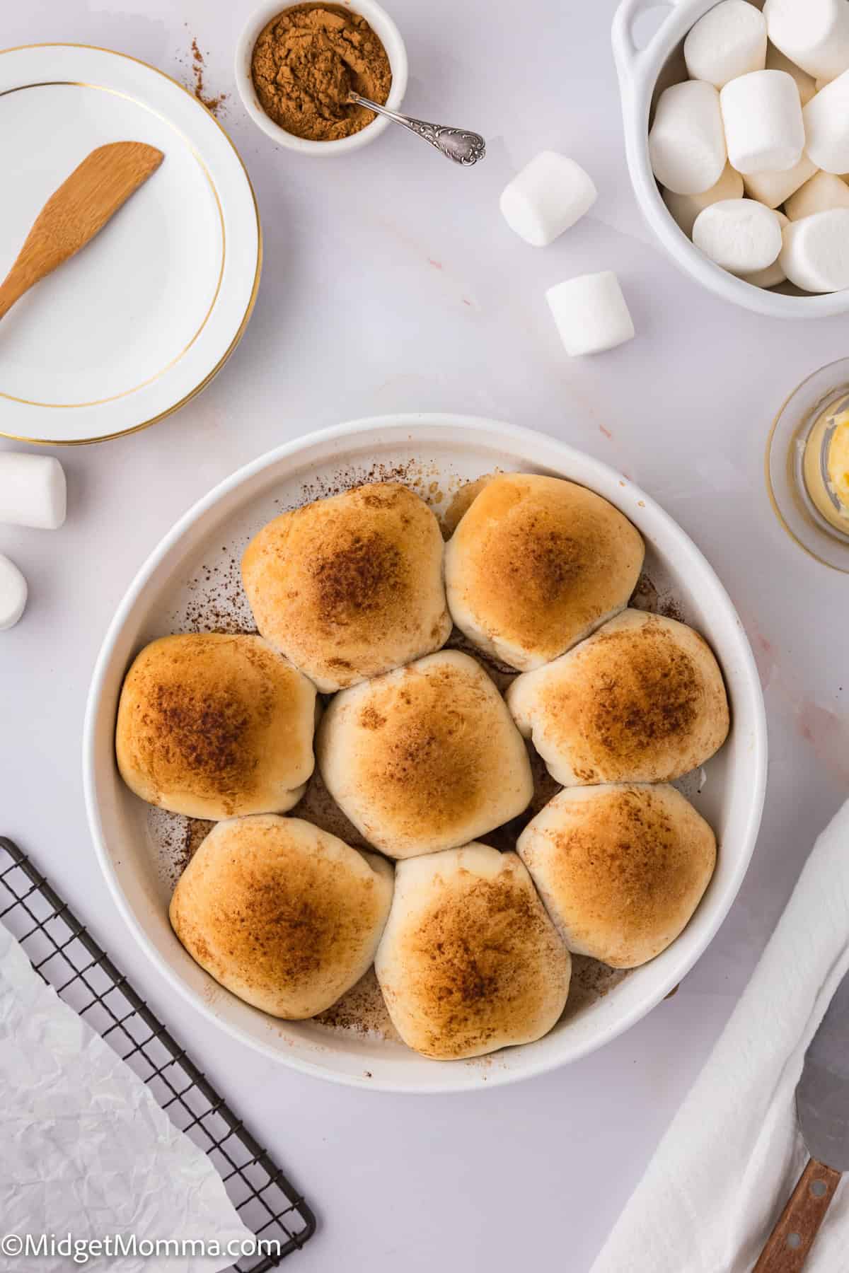 A round dish containing eight baked rolls, surrounded by marshmallows, a plate, a small bowl of brown sugar, and a rack on a white surface.