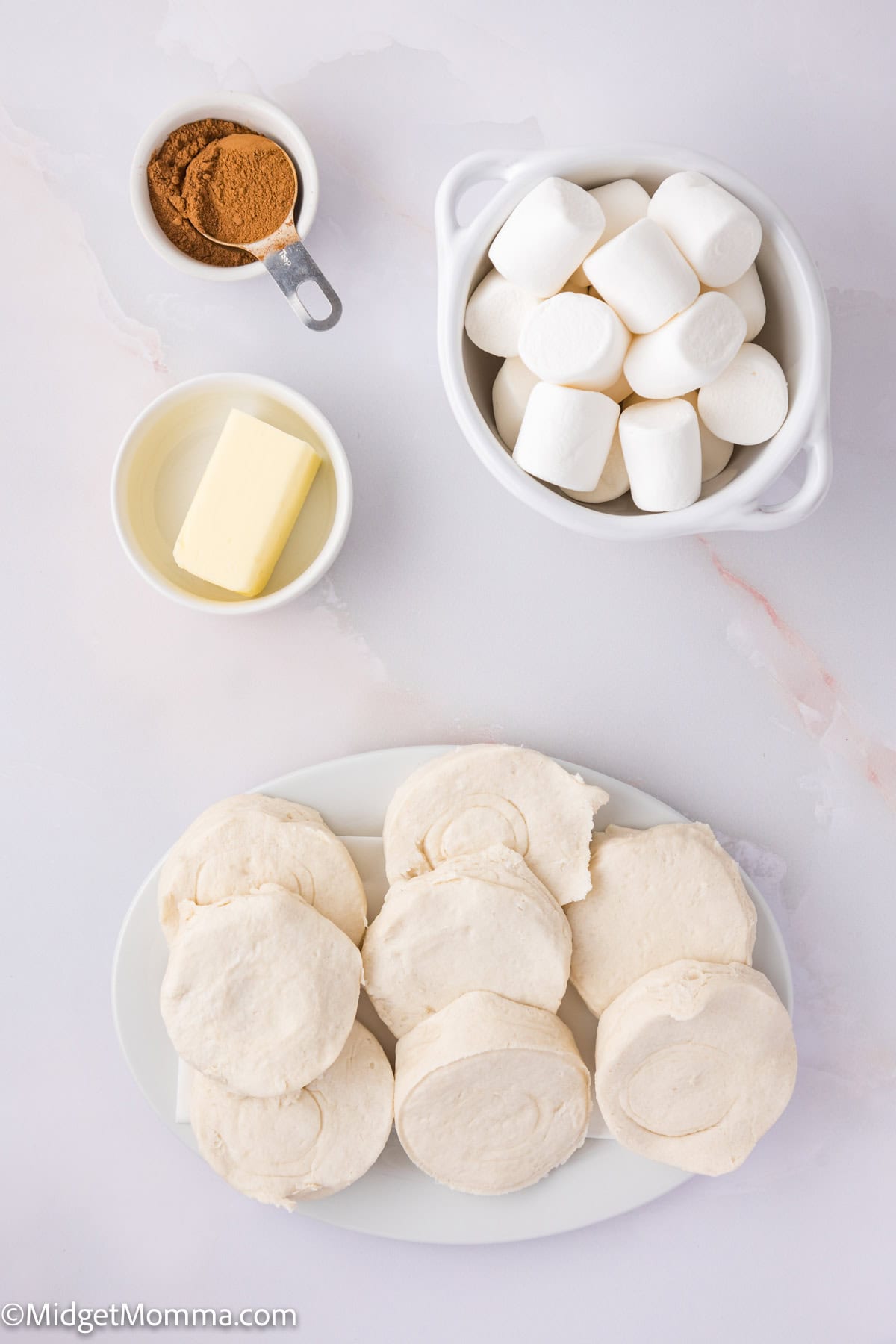 A white bowl of marshmallows, a small dish of butter, a spoonful of cinnamon, and several dough rounds on a plate arranged on a white surface.