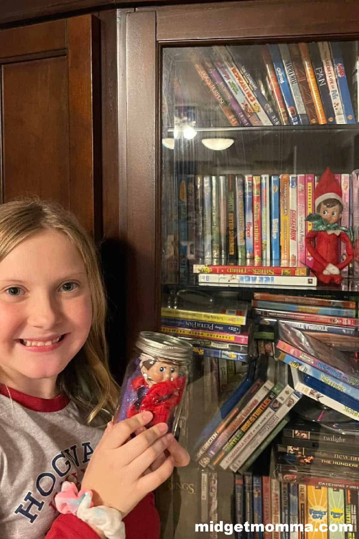A girl is holding a jar with elf on the shelf in it in front of a bookcase.