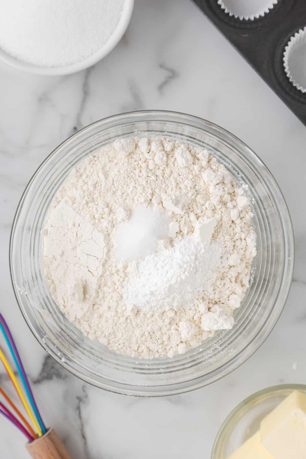 A bowl of flour, sugar, and other ry ingredients for mini blueberry muffins in a bowl on a marble countertop.